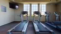 Located at 26900 Winchester Creek Ave in Murrieta, California, this single-floor fitness center is purpose-built for community wellness. The facility is accessed through a set of double glass doors at the entrance, which are framed by clean, neutral-toned walls and accented with a wall-mounted clock and sanitation station — details that speak to the facility's practical, well-maintained character.

The main workout area is generously proportioned and bathed in natural light from a bank of large, grid-paned windows along one wall. These windows frame views of palm trees and the surrounding landscape, creating an inviting atmosphere while working out. The flooring throughout consists of durable, interlocking rubberized tiles in a checkered pattern, designed to absorb impact and provide stability during exercise.

Cardio equipment is well-represented, with multiple treadmills and elliptical trainers positioned to take advantage of the natural light and outdoor views. A stationary bike and adjustable weight bench are also available for varied training options. A wall-length mirror spans one side of the room, visually expanding the space and aiding in form and technique. Ceiling fans are installed overhead to ensure comfortable air circulation during workouts, and wall-mounted flat-screen televisions provide entertainment. The overall ambiance is clean, functional, and welcoming.