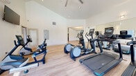 This comprehensive fitness center in Alameda, California is thoughtfully designed across a single floor, offering distinct zones for cardio training, strength work, and cycling. The space is characterized by striking vaulted ceilings that create an open, airy atmosphere, complemented by crisp white walls and light hardwood-style flooring throughout the cardio areas. Large windows allow generous natural light to fill the space while providing views of the surrounding community.

The cardio zone is well-appointed with multiple treadmills, elliptical trainers, a rowing machine, and upright and recumbent stationary bikes, all equipped with individual display screens. A dedicated spin studio features a row of cycling bikes with personal monitors, a ballet barre, and a full-length mirror wall — ideal for guided cycling and training sessions.

The strength training area transitions to a durable rubberized floor and is outfitted with a multi-functional cable machine, a full dumbbell rack, weight benches, kettlebells, stability balls, and resistance training accessories. Floor-to-ceiling mirrors amplify the sense of space throughout. A wall-mounted television, ceiling fans, and a mini-split HVAC unit ensure comfort during workouts. French doors in the strength zone open to an outdoor area, while an adjacent room with a billiards table hints at broader community amenities nearby.