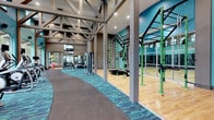 This expansive fitness center is designed to accommodate a wide range of workout styles and training needs, all within a single, thoughtfully designed space. The facility immediately impresses with its soaring vaulted ceilings, supported by exposed structural trusses that lend an open, airy industrial character to the environment. Teal-accented carpet flooring with rubber pathways defines distinct workout zones, adding both functionality and visual energy to the space. A dedicated functional training area features green powder-coated rig structures equipped with climbing ropes, gymnastic rings, and a rope climbing net, ideal for cross-training and bodyweight workouts. Adjacent to this zone, a wall of floor-to-ceiling mirrors amplifies the sense of space and supports form-focused training. The strength and cardio section offers a comprehensive selection of weight machines and free weights, arranged along a wide, easily navigable floor plan. A hardwood sports court, visible through the mirrored wall, extends the facility's versatility for court-based activities. Mounted televisions provide entertainment throughout the space. Natural light filters in through large windows, complementing the recessed lighting overhead and creating a bright, motivating atmosphere throughout the entire facility.