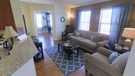 This single-floor residence offers a thoughtfully designed layout that balances comfort and functionality. The living area serves as the heart of the home, showcasing rich hardwood flooring that extends throughout the space. Large windows dressed with elegant draping curtains allow generous amounts of natural light to fill the room, creating a warm and inviting atmosphere. The living room is generously sized to accommodate comfortable seating arrangements, centered around a classic coffee table and anchored by a stylish patterned area rug that adds a decorative focal point to the space. Accent walls in warm earth tones provide a pleasing contrast to the lighter surrounding walls, adding depth and character to the interior design. Architectural details such as decorative column framing and crown molding contribute a refined, polished aesthetic. A dedicated office or study area is visible through an open doorway, complete with its own window for natural light, making it a practical and well-lit workspace. The open-concept flow between rooms enhances the sense of spaciousness throughout the home. Overall, the space presents a cohesive and comfortable interior with classic design elements and a welcoming ambiance.