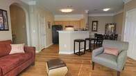 This single-floor residence offers an open-concept floor plan that integrates the kitchen, dining, and living spaces into one cohesive, flowing area. Warm hardwood flooring extends throughout, adding a sense of continuity and richness to the interior.

The kitchen is centrally positioned and features honey-toned wood cabinetry, black appliances including a full-size refrigerator, and a peninsula with a light-colored countertop. The peninsula serves as both a prep area and a casual dining spot, accommodating bar-height seating with sleek dark stools. Crown molding details add a refined architectural touch to the kitchen and surrounding spaces.

Adjacent to the kitchen, the dining area provides dedicated space for a table and chairs, while the living area is open and versatile, furnished with comfortable seating. The neutral, warm beige wall tones complement the flooring and cabinetry, creating a cohesive and inviting ambiance throughout.

Flush-mounted ceiling light fixtures provide consistent, even illumination across the main living space. White panel doors and crisp white baseboards contrast cleanly against the warm tones, lending a polished, well-maintained appearance. Overall, this residence presents a functional and comfortable layout suited to everyday living.