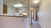 This single-floor residential unit offers a practical and well-organized layout designed for comfortable everyday living. The kitchen is a central feature of the space, equipped with white upper cabinetry that provides generous storage, a dark laminate countertop with a breakfast bar peninsula, a black electric range with an overhead exhaust hood, and a white refrigerator. Recessed under-cabinet lighting and a flush-mount ceiling fixture keep the kitchen bright and functional. The breakfast bar creates a natural transition between the kitchen and the adjoining living areas, making the space feel open and connected. A wide, carpeted hallway extends from the kitchen area, leading to the additional rooms of the unit. The hallway is well-lit with a ceiling-mounted light fixture and features warm wood-toned door trim that adds a subtle contrast to the neutral walls and light carpet throughout. Doors along the hallway provide access to what appears to be a bathroom and at least one additional room, suggesting a multi-room layout. The overall aesthetic is clean and neutral, with light-toned walls, textured ceilings, and carpeted flooring contributing to a cozy and understated interior atmosphere throughout the space.