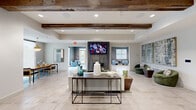Located at 1619 N Frazier Street in Conroe, Texas, this single-floor community clubhouse offers a well-appointed gathering space designed with a contemporary yet warm aesthetic. The main lounge area serves as the heart of the facility, featuring striking reclaimed wood ceiling beams that contrast beautifully against crisp white walls and large-format marble-look tile flooring. A sleek, tile-clad fireplace anchors the lounge and is complemented by a wall-mounted television, comfortable seating arrangements, and curated decorative accents. The open layout allows for generous circulation and a welcoming communal atmosphere. Adjacent to the lounge, a stylish bar-style kitchen counter with a chevron tile backsplash, pendant sconces, and bar seating provides an ideal space for casual socializing or community events. A dedicated meeting nook features a wooden dining table bathed in natural light from a large window, a statement brass pendant light, and built-in shelving. The game room, accessible from the main lounge, is equipped with a pool table and shuffleboard, with direct access to an outdoor pool area through sliding glass doors. Recessed lighting throughout enhances the modern ambiance, while large windows invite natural light across the entire space.
