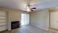 This single-story space centers around a comfortable living area finished with neutral-toned carpet that extends throughout the room. A fireplace positioned along one wall serves as a focal point, adding warmth and character to the space. A five-blade ceiling fan with an integrated light fixture is mounted to the textured ceiling, providing both air circulation and ambient lighting.

Natural light enters generously through a sliding glass door dressed with vertical blinds, which leads to an outdoor balcony with views of surrounding trees. This connection to the outdoors enhances the overall sense of openness in the room. The walls are painted in a warm, neutral beige tone that complements the carpeted flooring and contributes to a calm, understated ambiance.

A wall-mounted thermostat provides convenient climate control. White baseboards frame the perimeter of the room, offering a clean contrast against the neutral walls. The textured popcorn ceiling adds a classic architectural touch consistent throughout the space. Overall, the living area balances functionality and comfort with practical, well-considered details.