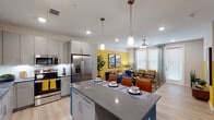 This well-appointed single-floor residence offers a thoughtfully designed open-concept layout that seamlessly connects the kitchen, dining, and living areas. The kitchen is a standout feature, boasting sleek grey flat-front cabinetry, a white subway tile backsplash, stainless steel appliances including a French door refrigerator, and a striking blue-accented island with dark quartz countertops and pendant lighting overhead. Bar seating at the island makes it an ideal space for casual dining and entertaining. The living area is bright and inviting, highlighted by a bold yellow accent wall, large windows that allow generous natural light, light wood-style flooring, and a ceiling fan. A private exterior door provides added convenience. In-unit stacked washer and dryer are tucked neatly within the unit. The primary bedroom features a sage green accent wall, warm wood furnishings, and ample natural light, while the secondary bedroom offers plush carpeting, a large window, and generous storage. The full bathroom includes a soaking tub, a white tile surround, grey cabinetry, dark quartz countertops, and a wide vanity mirror. Throughout the residence, a cohesive modern aesthetic is maintained with consistent finishes and vibrant design accents.