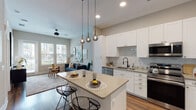 This single-floor residence offers a thoughtfully designed open-concept layout that seamlessly connects the kitchen and living areas. The kitchen is a standout feature, outfitted with crisp white shaker cabinetry, marble-style tile backsplash, quartz countertops, and a full suite of stainless steel appliances including a refrigerator, range, microwave, and dishwasher. A central island with seating provides additional prep space and a casual dining option, accented by sleek globe pendant lighting overhead. Warm hardwood-style flooring runs throughout, adding cohesion and a natural sense of warmth to the space. The living area is bathed in natural light from large windows with outdoor views, and is anchored by a ceiling fan and an inviting layout well-suited for relaxing or entertaining. The bedroom is a comfortable retreat featuring a window with sheer curtains, generous wall space, and rich dark hardwood flooring. The bathroom maintains the home's clean aesthetic with white cabinetry, a single-sink vanity with quartz countertop, an LED-lit mirror, and matte black fixtures. Neutral wall tones and contemporary styling create a cohesive, modern ambiance throughout the entire residence.