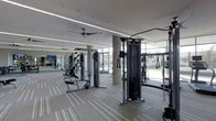 This contemporary fitness center offers a bright, open workout environment designed to inspire an active lifestyle. The space is defined by its expansive floor-to-ceiling windows that line the perimeter, flooding the interior with natural light and providing views of the surrounding outdoor landscape. Sleek linear LED lighting runs along the ceiling, complementing the natural light and ensuring the space is well-illuminated throughout the day and evening. The flooring features a modern striped carpet tile in neutral tones, adding a polished aesthetic while providing comfort underfoot. A ceiling fan is mounted overhead, contributing to air circulation across the workout floor. The facility is equipped with a comprehensive selection of strength training machines, including a cable cross machine and multi-function resistance units, catering to a wide range of fitness goals. Large mirrors are positioned strategically along the walls, enhancing the sense of space and allowing users to monitor their form during exercise. Structural columns are seamlessly integrated into the layout without impeding the flow of the floor plan. The overall ambiance is clean, modern, and professional, making this fitness center a well-appointed amenity suitable for a broad range of fitness enthusiasts.