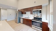 Located in the heart of downtown Worcester, MA, this two-bedroom, two-bathroom residence at 6 Chatham St offers a compelling blend of historic character and modern finishes across a generously sized single-floor layout. The combined kitchen and living area serves as the central hub of the home, featuring soaring ceilings, light hardwood flooring, and large windows that frame views of the surrounding cityscape. The kitchen is well-appointed with rich dark wood cabinetry, stainless steel appliances, a double-basin sink, and a center island with a clean white countertop — all beneath dramatic ceiling height that amplifies the sense of openness. The living room makes a striking impression with an exposed brick accent wall, a classic dark brick fireplace, plush carpeting, and a coffered ceiling detail with recessed lighting. The spacious primary bedroom features large windows offering urban views and a walk-in closet with built-in wire shelving on both sides. The second bedroom is similarly bright, with multiple windows and light hardwood floors. Both bathrooms are finished with white subway tile tub-and-shower surrounds, warm wood vanities, and neutral tile flooring. A dedicated closet with wire shelving adds further storage convenience throughout this well-designed urban residence.
