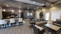 This single-floor communal space is thoughtfully designed to serve multiple functions within one open layout. The interior features a contemporary aesthetic with neutral gray tones, textured accent walls, and recessed lighting complemented by stylish pendant fixtures that add warmth to the environment.

The kitchen area is fully equipped with dark cabinetry, a stainless steel refrigerator, and a counter-height bar with seating, making it well-suited for catering or social gatherings. Adjacent to the kitchen, a compact dining area with a round table and chairs provides an intimate setting for smaller groups.

The lounge area is anchored by a large sectional sofa in a warm taupe tone, accented with teal throw pillows, and paired with a rustic coffee table. A ceiling-mounted projector indicates the space is designed to host screenings or presentations, adding a versatile entertainment dimension.

The flooring transitions between tile near the kitchen and patterned carpet in the lounge, helping to define each functional zone. Decorative framed artwork and a large potted plant contribute to the polished, welcoming ambiance throughout the space.