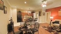This single-floor residence offers an open-concept layout that effortlessly connects the kitchen, dining, and living areas, creating a cohesive and functional living environment. The kitchen is well-equipped with warm wood cabinetry, dark appliances including a full-size refrigerator, recessed lighting, and a peninsula with bar-height seating — ideal for casual dining or entertaining. A pendant light fixture hangs above the peninsula, adding a decorative focal point to the culinary space. The dining area sits adjacent to the kitchen and features an elegant round table with ornate wrought-iron chairs, anchored beneath a ceiling fan with a light fixture that casts a warm ambient glow. Rich hardwood flooring flows throughout, complementing the neutral warm-toned walls and adding a sense of warmth and continuity across the open space. Recessed lighting throughout provides even illumination, while framed artwork adorns the walls, lending the space a curated, lived-in charm. A hallway visible from the main living area leads to additional rooms, suggesting a thoughtfully organized floor plan. Overall, the space conveys a comfortable, classic aesthetic with attention to detail in its finishes and furnishings.