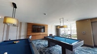 Located at 1600 Harbor Blvd in Weehawken, NJ, this single-floor recreational lounge is a well-appointed amenity space designed for leisure and social gathering. The room is anchored by two full-size pool tables with blue felt surfaces, each positioned beneath elegant pendant light fixtures that provide focused, atmospheric illumination. A warm wood-paneled feature wall serves as the focal point of the entertainment area, housing a large flat-screen television and built-in cabinetry — ideal for watching sports or hosting casual gatherings. The flooring throughout features a bold, geometric-patterned carpet in deep blues, blacks, and gold tones, lending the space a sophisticated, contemporary character. Natural light filters in generously through floor-to-ceiling windows, offering views of the surrounding outdoor area and creating an airy, open ambiance. Near the windows, a bistro-style high-top table with curved modern chairs provides an intimate seating nook. The entry area is accented with a decorative gold console table, a framed mirror, and floral arrangements, establishing an upscale first impression. Abstract artwork adorns the walls, complementing the modern aesthetic. The overall design balances energetic recreational functionality with refined interior detailing, making it a versatile and inviting communal space.
