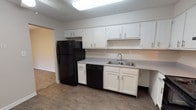 Regal Park is a single-floor residence designed with comfort and practicality in mind. The kitchen is outfitted with white cabinetry providing ample storage, a double-basin stainless steel sink, grey laminate countertops, and black appliances including a full-size refrigerator and dishwasher. Wood-look flooring runs throughout the kitchen, while a bright overhead light fixture keeps the space well illuminated.

The living room is generously proportioned, featuring plush carpeting, neutral-toned walls with a standout blue accent wall, and a large sliding glass door that floods the room with natural light and offers a view of the outdoors. A paneled front entry door provides a welcoming entrance to the space.

Adjacent to the kitchen, the dining area benefits from a stylish pendant light fixture and carpeted floors, with a direct sightline into the kitchen for a connected, open feel.

The bedrooms are comfortable and well-lit, each featuring soft carpeting, ceiling fans with light fixtures, and windows with blinds that allow for natural light. Textured ceilings are consistent throughout the unit, adding a classic residential character. The overall aesthetic is clean, neutral, and move-in ready, offering a practical and inviting living environment.