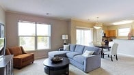 This single-floor space offers a well-designed open-concept layout that seamlessly connects the living and dining areas. The living room is anchored by a spacious gray sectional sofa, complemented by a round dark-wood coffee table and soft, neutral-toned carpeting underfoot. Warm taupe walls create a cozy yet sophisticated atmosphere throughout.

Natural light pours in generously through multiple large windows, offering pleasant outdoor views of mature trees and the surrounding landscape. The windows are dressed with clean, simple treatments that enhance the bright, airy feel of the room.

The adjacent dining area is visible from the living space, furnished with a dark dining table and chairs beneath a classic pendant light fixture, creating a cohesive and inviting gathering space. A white paneled door adds a traditional architectural touch, while crown molding along the ceiling contributes an elegant finishing detail.

Overall, this space strikes a balance between comfort and style, with a neutral color palette and thoughtful furnishings that give it a welcoming, livable quality. The open flow between rooms makes the space feel expansive and functional, ideal for both everyday living and entertaining.