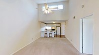 Located at 100 Lake Clair Place in Fayetteville, North Carolina, this single-floor residence offers a thoughtfully designed open-concept layout that maximizes both space and functionality. The living area immediately impresses with its dramatically vaulted ceilings, which lend an airy, expansive feel to the room. A ceiling fan with a light fixture is centrally mounted at the peak, providing both airflow and illumination. A high clerestory window near the top of the wall allows natural light to filter into the space, brightening the neutral-toned interior throughout the day.

Soft, light-colored carpeting covers the living area floor, contributing to a clean and comfortable atmosphere. The space transitions seamlessly into the kitchen, which features white cabinetry, white appliances including a refrigerator and range, and a peninsula-style breakfast bar with two bar stools — ideal for casual meals or entertaining. The kitchen area transitions to a harder flooring surface, clearly delineating it from the carpeted living space. The overall color palette throughout the home is neutral and understated, with white and beige tones creating a bright, welcoming environment throughout this single-level residence.