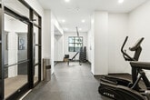Located in Missoula, Montana, this compact yet fully outfitted gym delivers a comprehensive fitness experience within a thoughtfully designed space. The room features a clean, modern aesthetic with crisp white walls, recessed lighting, and durable rubber-speckled flooring that provides both traction and comfort during workouts.

The facility is divided into distinct functional zones. A strength training area is anchored by a full dumbbell rack with a wide range of weights, an adjustable workout bench, and a suspended TRX suspension training system, ideal for functional fitness. A large floor-to-ceiling mirror expands the sense of space while allowing users to monitor their form. Natural light filters in through multiple windows, offering views of the surrounding Missoula neighborhood.

The cardio zone is equipped with a treadmill and an elliptical trainer, both positioned for practical use. A wall-mounted flat-screen TV adds entertainment for longer sessions. A stability ball rounds out the available equipment for core and balance training. Storage is provided by a wire shelving unit near the entrance.

The gym is accessed through a stylish black-framed glass door, adding an industrial-modern touch to the overall design. Abstract framed artwork on the walls contributes a polished, intentional aesthetic. This space is well-suited for a variety of workout routines and offers a focused, distraction-free environment for residents seeking convenient on-site fitness amenities.