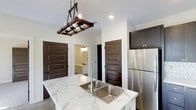 This single-floor space showcases a thoughtfully designed interior where modern finishes and rustic touches come together seamlessly. The kitchen serves as a central highlight, anchored by a substantial island topped with striking marble-look countertops featuring bold veining that adds both elegance and visual interest. A deep double-basin stainless steel sink is set into the island, complemented by a sleek modern faucet. Overhead, a distinctive wood-and-metal industrial chandelier with exposed bulbs adds a warm, rustic character to the space. Dark espresso-toned cabinetry lines the walls, providing rich contrast against the crisp white walls and ceiling, while a full-size stainless steel refrigerator completes the functional kitchen suite. Recessed lighting throughout ensures the space is well-lit and inviting. Rich, dark panel-style doors lead to adjoining rooms, maintaining a consistent design language throughout. A glimpse into a connected room reveals additional cabinetry and a sink, suggesting a well-appointed bathroom nearby. The overall ambiance balances contemporary functionality with warm, artisanal design details, creating a space that feels both refined and welcoming.