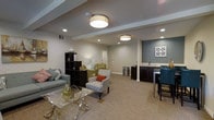 This thoughtfully designed single-floor space offers a seamless blend of comfort and functionality. The main living area is anchored by a light-toned carpet that runs throughout, complementing the neutral color palette of the walls and ceiling. Recessed lighting, paired with elegant drum pendant fixtures, provides warm, layered illumination across the entire space.

The lounge area is furnished with a mix of seating options, including a plush sofa adorned with accent pillows, a patterned accent chair, and additional side chairs, all arranged around a sleek glass-top coffee table. A dark-toned open shelving unit serves as both a functional divider and a display feature between seating zones.

On one end of the space, an accent wall in a deep teal-gray hue adds a striking visual contrast, framing a built-in wet bar area. The bar features dark cabinetry, a countertop with a sink, and bar-height seating, making it an ideal spot for entertaining. A decorative wall art piece above the bar adds a refined finishing touch.

The space also includes a secondary seating nook with a pair of armchairs, offering a quieter retreat within the open layout. A doorway at the far end provides additional access, contributing to the overall sense of flow and openness throughout the space.