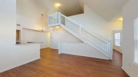 This two-floor residence is defined by its dramatic vaulted ceilings that soar above the open living area, creating an expansive, airy atmosphere filled with natural light from multiple windows, including a distinctive circular accent window. Warm hardwood-style flooring flows throughout the main living spaces on both levels, adding cohesion and a welcoming feel.

The living area serves as the heart of the home, anchored by a striking white staircase with classic baluster railings that leads to an open upper-floor landing overlooking the space below. A chandelier adds a touch of elegance to the lower level, while ceiling fans on both floors provide practical comfort.

The galley-style kitchen is well-appointed with honey-toned wood cabinetry, granite-look countertops, and a full suite of black appliances including a refrigerator, electric range, microwave, and dishwasher. A window at the end of the kitchen brings in natural light.

The bathroom features warm oak cabinetry with a granite-look countertop and single-basin sink, complemented by tile flooring and a tub-shower combination with decorative tile accents. Additional rooms include generously sized bedrooms with vaulted ceilings, ceiling fans, and ample natural light, offering a bright and comfortable environment throughout.