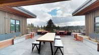 This rooftop terrace offers an inviting open-air gathering space defined by a clean, contemporary design. The large-format stone tile flooring provides a durable and polished foundation, while a partial overhead canopy with warm wood-tone ceiling detail offers limited shelter without compromising the openness of the space. Flanking structures clad in vertical wood siding frame the terrace on either side, contributing a natural, modern aesthetic.

The seating arrangement is well-considered and varied. A central picnic-style table with a light wood slat top and black steel base is accompanied by matching benches, serving as the primary communal gathering point. Surrounding the perimeter, smaller bistro-style tables paired with bold red chairs and matching red pedestal bases add a pop of color and provide more intimate seating options. Built-in raised planters along the perimeter railing add greenery and definition to the space.

The terrace's most notable feature is its unobstructed panoramic view, encompassing mature evergreen trees, distant forested hillsides, and a sweeping open sky — making it a standout space for outdoor gatherings and relaxation.