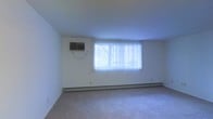 This single-floor space offers a clean and functional layout with a straightforward, practical design. The main room is spacious and open, featuring wall-to-wall carpeting that adds warmth and softness underfoot. A large window dressed with vertical blinds serves as the focal point of the room, allowing abundant natural light to fill the interior and creating a bright, airy atmosphere. A baseboard heating unit runs along the base of the exterior wall beneath the window, providing efficient climate control throughout the colder months. Complementing this, a wall-mounted air conditioning unit ensures year-round comfort. The walls are finished in a neutral white tone, lending the space a clean and versatile backdrop that is well-suited to a variety of furnishing styles. The room's uncluttered design and open floor plan provide flexibility in how the space can be arranged and utilized. Overall, this space presents a simple, well-maintained environment with essential features in place to support comfortable, everyday living.