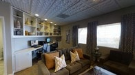 This thoughtfully designed single-floor space seamlessly combines a relaxed living area with a productive home office environment. The room's most striking architectural feature is its ornate embossed ceiling tiles, which lend a distinctive, textured elegance overhead, complemented by recessed lighting that creates a warm and inviting atmosphere.

A generously sized tufted sofa anchors the lounge area, dressed with richly patterned accent pillows in warm amber and gold tones. A sleek glass-top side table and a geometric metallic accent piece add contemporary flair to the seating zone.

Along one wall, a built-in workstation accommodates dual computer monitors, flanked by ergonomic office chairs — making the space equally suited for professional productivity. Above the desk, floor-to-ceiling open shelving in crisp white displays an artful collection of decorative vases, books, and curated accents, adding both function and visual depth.

The walls are finished in a neutral warm gray, providing a cohesive backdrop that ties together both zones. Natural light filters in through windows dressed with bold chevron-patterned curtains, softening the overall ambiance. Dark hardwood-style flooring grounds the space and enhances the sophisticated, versatile character of this well-appointed interior.