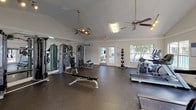 This fitness center is thoughtfully designed to support a comprehensive workout experience within a bright, open, single-floor layout. The space features vaulted ceilings that lend an airy, expansive feel, complemented by multiple ceiling fans and a combination of track lighting and overhead fluorescent fixtures that ensure the room remains well-lit throughout the day. Large windows and glass-paned French doors line the walls, inviting abundant natural light and offering views of the surrounding outdoor greenery, creating an energizing atmosphere for exercise. The flooring throughout is a durable rubber surface suited for both heavy equipment and high-intensity activity. Full-length mirrors span one wall, visually enlarging the space and allowing users to monitor their form during workouts. The strength training area is outfitted with multi-station cable machines, a weight bench, and various resistance equipment, while the cardio zone features treadmills, an elliptical trainer, and additional aerobic machines. A wall-mounted flat-screen television adds to the amenity, offering entertainment during cardio sessions. A medicine ball rack and dedicated stretching areas round out the offerings. The clean, neutral color palette of white walls and gray flooring gives the fitness center a modern, professional appearance.