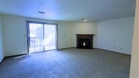 This single-floor space offers a welcoming and functional layout centered around a comfortable living area. The room is anchored by a classic wood-mantled fireplace, which serves as a striking focal point and adds warmth and character to the space. Smooth, neutral-toned walls provide a clean, versatile backdrop throughout.

Plush wall-to-wall carpeting covers the living area floor, contributing to a cozy and comfortable atmosphere underfoot. A large sliding glass door fitted with vertical blinds lines one wall, allowing an abundance of natural light to flood the interior and offering direct access to the outdoors. The combination of the bright, open feel from the expansive glazing and the warmth of the fireplace creates a balanced and inviting ambiance.

The ceiling is smooth and painted in a cool neutral tone, complementing the overall light and airy feel of the space. Ceiling-mounted ventilation registers indicate a central HVAC system for year-round climate control. The single-floor layout ensures easy accessibility and a straightforward flow throughout the interior. Overall, the space presents a clean, move-in ready environment with timeless features suited to a variety of furnishing and design preferences.