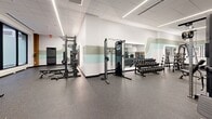 Located at 608 Second Ave S in downtown Minneapolis, this fitness center offers a generously sized, single-floor exercise facility designed to support a full range of workout routines. The space is defined by a clean, modern aesthetic, featuring durable speckled rubber flooring, a drop tile ceiling with bright linear LED lighting, and a distinctive wavy multi-tone mural that runs along the walls in soft shades of sage green and aqua, lending the space a polished, energetic atmosphere.

The main exercise room is comprehensively equipped, with a dedicated cardio zone featuring a row of treadmills and stair climbers, each fitted with individual digital screens. Strength training is well represented through selectorized cable and resistance machines, a power rack with barbells and weight plates, and a full dumbbell rack spanning a wide range of weights. Spin bikes and a pull-up and dip station occupy a windowed alcove, allowing natural light to filter into the space. Full-length mirrors are strategically placed throughout to maximize sightlines.

A hallway entrance welcomes visitors with framed black-and-white sports photography, setting an athletic tone from arrival. Wood-accented columns and locker storage add warmth to the otherwise crisp, white interior. The overall layout is open and well-organized, providing ample floor space for movement between equipment zones.