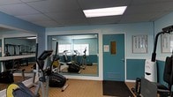 This single-level fitness room offers a practical and functional workout space equipped with essential cardio machines, including stationary bikes and an elliptical trainer. The room is finished with teal-painted walls accented by white trim, creating a clean and energizing atmosphere conducive to exercise.

A large wall mirror spans a significant portion of one wall, framed in matching teal, which visually expands the space and allows users to monitor their form during workouts. The drop ceiling features a large flush-mounted fluorescent light panel that provides bright, even illumination throughout the room.

The flooring consists of a neutral patterned carpet, which adds a degree of comfort underfoot, while a rubber entry mat near the door helps maintain cleanliness. A dedicated exit door, clearly marked with a no-smoking sign, provides convenient access. A framed informational board is mounted on the wall, likely detailing facility rules or equipment guidelines. A floor-level ventilation grille ensures adequate air circulation, contributing to a comfortable environment during use.