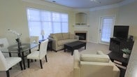 This single-floor residence offers an open and functional layout centered around a well-appointed living area. The space is finished with neutral-toned carpeting and warm beige walls, creating a calm and cohesive atmosphere throughout. Crown molding runs along the ceiling, adding a subtle architectural refinement to the interior.

The living area serves as the focal point of the space, anchored by a corner fireplace with a classic arched surround and a clean white mantelpiece. Large windows fitted with horizontal blinds line the walls, allowing generous amounts of natural light to fill the room while maintaining privacy. A glass-panel door provides direct access to an outdoor area, further enhancing the sense of openness.

The seating arrangement includes a neutral sofa and accompanying armchairs, complemented by a dark coffee table that provides contrast against the light-toned furnishings. Adjacent to the living area, a glass-top dining table with white upholstered chairs offers a dedicated space for meals. The overall ambiance is bright, airy, and understated, with a design sensibility that prioritizes comfort and practicality.