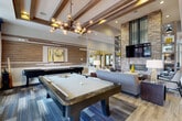 Located at 1111 Oakley Industrial Blvd in Fairburn, GA, this single-floor community clubhouse offers a well-appointed amenity space designed with modern, upscale finishes throughout. The grand entry lobby sets an impressive tone with soaring double-height ceilings, bold geometric wall treatments in yellow and gray, floor-to-ceiling windows that flood the space with natural light, and light wood-look flooring that carries throughout the facility. Exposed wood beam ceilings add warmth and architectural character to the main gathering areas. The expansive lounge features a dramatic floor-to-ceiling brick fireplace, a wall-mounted TV, built-in shelving, comfortable seating arrangements, and a full-size billiards table, making it an ideal space for both relaxation and recreation. A cozy lounge nook nearby offers built-in bench seating, stylish pendant lighting, and patterned carpeting for a more intimate atmosphere. The conference and dining room provides a large wood table with leather seating, abundant natural light from oversized windows with views of the outdoors, and elegant pendant lighting. The kitchenette is outfitted with warm wood cabinetry, open floating shelves with decorative accents, stainless steel appliances including a microwave and beverage cooler, a coffee station, and a sink — all set against bold navy blue walls with textured tile backsplash.