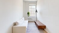 Located at 1228 Spruce Street in Philadelphia's vibrant Washington Square West neighborhood, this one-bedroom, one-bathroom unit offers a thoughtfully organized single-floor layout. Light wood flooring runs throughout the main living areas, complementing a clean, all-white color palette that keeps the space feeling open and airy.

A narrow hallway greets you at the entry, leading past the kitchenette — fitted with white cabinetry and a compact countertop workspace — and into the living room. The living room is the social heart of the unit, featuring a large arched window that frames views of the surrounding Philadelphia cityscape and draws in natural light. The space accommodates a sectional sofa, a low-profile media console, and an area rug.

The bedroom is an intimate, efficiently arranged room with a window fitted with an in-unit air conditioning unit, providing both ventilation and climate control. The room offers just enough space for a full-sized bed and a small side table.

The bathroom features neutral tile flooring, white walls, and a simple, functional design. A dedicated closet provides additional storage. Throughout the unit, the minimalist aesthetic, consistent finishes, and natural light create a calm, practical living environment well-suited for urban living in Center City Philadelphia.
