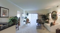 This single-floor space centers around a comfortably sized living room that draws immediate attention with its large sliding glass doors dressed in vertical blinds, allowing an abundance of natural light to fill the room. Crown molding trims the smooth, light-colored ceiling, adding a touch of classic architectural detail throughout the space. The neutral wall-to-wall carpeting contributes to a warm, cohesive aesthetic.

The living room is thoughtfully arranged with a mix of traditional furnishings, including an ornate accent chair with floral upholstery and a glass-top coffee table that reflects the room's ambient light. A plush sofa sits to one side, complementing the overall seating arrangement. Lush indoor plants are positioned throughout, adding vibrant greenery that enlivens the interior.

Wall art, including a framed piece near the sliding doors and a vertical framed print on an adjacent wall, adds a curated, lived-in character. A doorway visible from the living area hints at additional rooms beyond, suggesting a practical and well-connected layout. Overall, the space conveys a pleasant, classic residential ambiance with an emphasis on natural light and greenery.