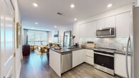 Located at the iconic 6200 Sunset Blvd in the heart of Hollywood, Los Angeles, this modern single-floor residence combines contemporary design with functional living. The open-concept layout seamlessly connects the kitchen, dining, and living areas, creating a bright and cohesive space bathed in natural light from large windows with horizontal blinds.

The kitchen is a standout feature, equipped with sleek white flat-panel cabinetry extending to the ceiling, dark stone countertops, a marble-style tile backsplash, and a full suite of stainless steel appliances, including a range, microwave, dishwasher, and French door refrigerator. A peninsula with bar seating bridges the kitchen and dining area, ideal for casual or formal gatherings.

The dining area accommodates a round wood dining table and connects fluidly to a comfortable living space. Wide-plank wood-look flooring runs throughout the main living areas, adding warmth and continuity.

The bedroom is generously proportioned, featuring a ceiling fan, access to a private outdoor area through glass doors, and an en-suite bathroom. The bathroom is finished with large-format tile flooring, a floating vanity with dark countertop, and a soaking tub with marble-style tile surround. Recessed lighting and a clean, neutral palette complete the refined aesthetic of this well-appointed Hollywood residence.