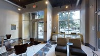 This single-floor commercial space presents a sleek, contemporary lobby designed to make a strong first impression. The entrance is anchored by a set of glass double doors framed by large, floor-to-ceiling windows that flood the interior with abundant natural light and offer clear views of the surrounding streetscape and mature trees outside. The high ceilings, finished in a dark tone, create a striking contrast against the light-colored walls and pale tile flooring, which transitions into a reflective metallic inlay near the entrance — a distinctive architectural detail that adds visual interest underfoot. Overhead, a series of pendant lights hang at varying heights, contributing to the modern aesthetic while providing warm, focused illumination throughout the space. The seating area is well-appointed with upholstered lounge chairs and small round accent tables, arranged to encourage both individual use and casual conversation. A clean white cube ottoman adds a minimalist touch to the lounge zone. Framed artwork on the walls and a sculptural decorative element near the window ledge lend the space personality and polish. Overall, the lobby balances open, airy proportions with thoughtfully curated furnishings to create a welcoming and professional atmosphere.