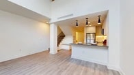 This well-designed two-bedroom, two-bathroom residence spans two floors, offering a thoughtful layout that balances open communal living with private upper-floor retreats. The main floor features a combined kitchen and living area distinguished by soaring double-height ceilings that amplify the sense of space and airiness. Light hardwood-style flooring flows throughout, complementing the clean white walls and modern aesthetic. A sleek staircase with classic wood balusters connects the two levels seamlessly.

The contemporary kitchen is a standout feature, outfitted with white cabinetry, a herringbone tile backsplash, stainless steel appliances including a refrigerator, range, and over-the-range microwave, and a peninsula with a stone countertop illuminated by striking pendant lights. The peninsula offers casual seating and additional prep space.

Upstairs, the generously sized primary bedroom provides ample room for furnishings, while the second bedroom offers a comfortable and versatile space. Both bathrooms are finished in a clean, minimalist style with large-format tile flooring, wood-tone vanities, white quartz countertops, and modern fixtures. The upper-level full bathroom includes a soaking tub and tiled shower surround. A dedicated closet completes the upper floor's storage offerings.