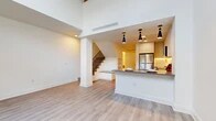 This well-designed two-bedroom, two-bathroom residence spans two floors, offering a thoughtful layout that balances open communal living with private upper-floor retreats. The main floor features a combined kitchen and living area distinguished by soaring double-height ceilings that amplify the sense of space and airiness. Light hardwood-style flooring flows throughout, complementing the clean white walls and modern aesthetic. A sleek staircase with classic wood balusters connects the two levels seamlessly.

The contemporary kitchen is a standout feature, outfitted with white cabinetry, a herringbone tile backsplash, stainless steel appliances including a refrigerator, range, and over-the-range microwave, and a peninsula with a stone countertop illuminated by striking pendant lights. The peninsula offers casual seating and additional prep space.

Upstairs, the generously sized primary bedroom provides ample room for furnishings, while the second bedroom offers a comfortable and versatile space. Both bathrooms are finished in a clean, minimalist style with large-format tile flooring, wood-tone vanities, white quartz countertops, and modern fixtures. The upper-level full bathroom includes a soaking tub and tiled shower surround. A dedicated closet completes the upper floor's storage offerings.