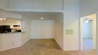 This single-floor space presents a clean, open layout finished in neutral white tones throughout. The main living area features wall-to-wall carpeting in a warm beige hue, providing a comfortable foundation that complements the light-colored walls. Recessed lighting in the ceiling offers functional overhead illumination, contributing to a bright and even atmosphere within the space.

A defining architectural feature is the partial wall with a lowered soffit that separates the living area from the adjacent kitchen zone, creating a semi-open connection between the two spaces. The kitchen area, partially visible to the left, includes upper white cabinetry and a dark countertop, suggesting practical functionality. A built-in panel — likely a utility or electrical access panel — is mounted on the right wall, adding a practical element to the space.

The white baseboards provide a clean contrast against the carpeted floor, while the overall minimalist finish of the walls allows for versatile interior customization. The layout feels open and adaptable, making it well-suited for a range of furnishing configurations. The space presents a neutral, move-in-ready aesthetic with a straightforward and functional design.