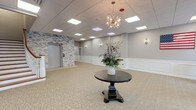 This refined lobby space presents a thoughtfully designed interior that balances classic elegance with contemporary style. The open, generously proportioned floor plan is anchored by a striking dark wood pedestal table topped with a decorative floral arrangement, beneath a sculptural sputnik chandelier that serves as a visual centerpiece. The drop ceiling is fitted with recessed panel lighting, ensuring the space is evenly and warmly illuminated throughout.

The walls are finished in a soft gray palette and accented with white wainscoting panels, lending a polished, formal character to the interior. Textured stacked-stone accent walls provide an organic contrast, framing both the elevator and the main entry door. A white-railed staircase with wood treads rises gracefully along one wall, offering an architectural focal point and access to upper levels.

A seating area near oversized, dark-framed floor-to-ceiling windows invites natural light into the space while offering views of the surrounding brick exterior streetscape. Tufted leather armchairs add a refined, professional touch to this corner. Near the entryway, a glass-panel door set within a stacked-stone surround provides an inviting transition from the exterior. An American flag displayed on the wall adds a patriotic accent. The plush neutral carpeting throughout completes the welcoming and professional ambiance of this well-appointed space.
