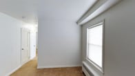 This single-floor unit offers a clean, straightforward layout designed for comfortable everyday living. The living and bedroom areas feature warm beige carpeting throughout, crisp white walls, and structural beam detailing along the ceilings that add a subtle architectural character. Multiple windows fitted with blinds allow natural light to fill the rooms, while baseboard radiator units provide efficient heating. The bedroom includes dual closets with open shelving and hanging rods, offering generous built-in storage. A hallway connects the rooms with white paneled doors, maintaining a cohesive and tidy aesthetic throughout. The kitchen is outfitted with warm honey-toned oak cabinetry, both upper and lower, paired with stone-patterned laminate countertops and a stainless steel sink. Light blue-tinted walls in the kitchen create a subtle contrast against the wood tones. The bathroom presents a classic design with a full-size soaking tub and overhead shower, white tile surround, and a distinctive black accent tile border that adds a timeless touch. A frosted window in the bathroom allows for natural light while maintaining privacy. Overall, the space presents a functional and well-maintained interior with traditional detailing throughout.
