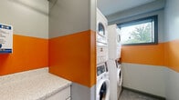 This compact shared laundry room is designed for efficiency and functionality, making the most of its modest footprint. The space is immediately distinguished by its bold two-tone wall treatment — a vibrant orange band runs horizontally across the mid-section of each wall, contrasting sharply with the cool light gray above and below, creating a lively and energetic atmosphere. The polished concrete floors add an industrial character while remaining easy to maintain. Multiple stacked washer and dryer units line the walls, maximizing the available floor space and providing ample laundry capacity. A built-in white cabinetry unit with a speckled stone-look countertop offers a dedicated folding and sorting surface, keeping the workflow organized. A payment terminal mounted above the counter indicates coin-free, card-based access for user convenience. Safety is addressed with a clearly visible fire extinguisher and alarm near the entry. Natural light filters into the room through a framed window, offering a pleasant outdoor view of surrounding greenery. The gray-trimmed entry door and clean sightlines throughout the room contribute to a tidy, well-organized shared amenity space that balances practicality with a distinctive visual design.