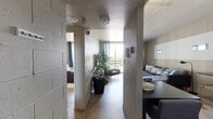 Located at 303 E Congress St in the heart of downtown Tucson, Arizona, this single-floor unit offers an urban industrial aesthetic defined by exposed concrete block walls, polished concrete floors, and a functional, open layout. A central hallway connects the living spaces, creating an efficient flow throughout.

The living area is a standout feature, anchored by large floor-to-ceiling sliding glass doors that open to a private balcony with sweeping views of the Tucson skyline and palm tree-lined streetscape. Teal drape panels frame the doors, softening the industrial materiality. The space comfortably accommodates a seating arrangement and media setup against the textured block walls.

The kitchen is equipped with a full-size refrigerator, electric range, dishwasher, and sink, complemented by open shelving and a compact dining area with a table and chairs.

The bedroom benefits from its own direct balcony access through a sliding glass door, offering abundant natural light and outdoor views. Built-in closet storage lines one wall, providing practical organization.

The bathroom features a modern white vanity with under-cabinet storage, an overhead mirror, and clean, minimal finishes. Throughout the unit, cage-style industrial light fixtures reinforce the contemporary urban character of the space.