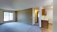 This single-floor space features an open-concept layout that connects the main living area to the kitchen and additional rooms. The living area is finished with neutral-toned carpeting and painted walls in a warm beige, creating a understated, functional atmosphere. A textured ceiling adds subtle dimension overhead, while a window along one wall allows natural light to brighten the space.

A structural column and partial wall define the boundary between the living area and the kitchen zone without fully closing off the floor plan. The kitchen is visible to the right, where warm wood-toned cabinetry provides storage and a countertop peninsula serves as a transitional element between the two areas. The kitchen section features hard flooring, contrasting with the carpeted living space.

Toward the back of the unit, a hallway leads to additional rooms, including what appears to be a bedroom or storage area behind a wood-paneled door. The overall layout is practical and straightforward, with a neutral color palette throughout that provides a versatile backdrop. The space offers a modest but functional design suited for everyday living.