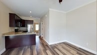 This single-floor cottage at The Cottages offers a thoughtfully designed open-concept layout that seamlessly connects the kitchen and living areas. The kitchen is well-appointed with rich dark cabinetry, granite countertops, a stainless steel refrigerator, and a central island with a built-in sink — providing ample workspace and storage. The warm hardwood-style flooring flows throughout the space, adding a sense of continuity and warmth. Crown molding accents the ceilings, lending an elevated, refined touch to the interior. Neutral, light-toned walls create a bright and airy atmosphere, enhancing the sense of openness throughout the home. Recessed lighting illuminates the kitchen area, complemented by a ceiling fan that adds both function and comfort to the living space. A doorway leads to an additional room, offering natural light and connectivity between areas. The clean, well-maintained finishes and practical layout make this cottage a comfortable and functional single-floor residence suited for easy everyday living.