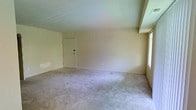 This single-floor residence offers a clean, functional layout designed for comfortable everyday living. The main living area is a standout feature of the space, presenting generous square footage with plush wall-to-wall carpeting in a neutral tone that lends a warm, understated feel underfoot. The walls are finished in a soft, light beige palette throughout, contributing to a bright and cohesive interior atmosphere.

Natural light plays an important role in the ambiance of the living area, with a large window along one wall allowing daylight to fill the room and create an open, airy feel. The ceiling is smooth and uniform, reinforcing the clean lines of the space.

An interior door is visible within the main room, providing access to additional areas of the residence and suggesting a practical, connected floor plan. Electrical outlets are conveniently positioned along the baseboards, offering flexibility for furniture arrangement and everyday use.

Overall, this space presents a neutral, move-in ready interior with a simple and versatile design that can be easily personalized. The combination of soft carpeting, ample natural light, and a well-proportioned main living area makes it a functional and adaptable single-floor residence suitable for a variety of lifestyles.