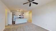 This single-story residence features three bedrooms and two bathrooms within a thoughtfully arranged floor plan. The open-concept living and kitchen area serves as the heart of the home, creating a cohesive and functional shared space. The kitchen is equipped with stainless steel appliances, including a refrigerator and range, and is accented by light wood cabinetry with a tile backsplash and dark stone countertops. A central kitchen island provides additional workspace and casual seating, while pendant lighting above the island adds a decorative touch alongside recessed ceiling lights. The living area features neutral wall-to-wall carpeting that contributes to a warm, comfortable atmosphere, and a ceiling fan overhead promotes air circulation throughout the space. The clean, neutral color palette of white walls and light finishes gives the home a bright, modern feel. The layout flows openly between the kitchen and living space, making it well-suited for everyday living and entertaining. The overall design emphasizes practicality and comfort, with consistent finishes throughout that create a cohesive aesthetic.