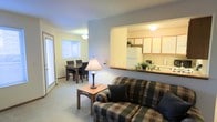 This single-floor unit offers an open-concept layout that seamlessly connects the living room, dining area, and kitchen. The living room is anchored by a plaid sofa with coordinating throw pillows and a warm wooden side table with a classic table lamp, creating a comfortable and inviting atmosphere. Neutral-toned carpet runs throughout, adding softness underfoot.

A pass-through counter opens directly into the kitchen, which is equipped with light wood cabinetry, a white refrigerator, and ample counter space for meal preparation. The open sightline between these areas enhances the sense of space and promotes ease of movement throughout.

Adjacent to the living area, the dining space accommodates a dark wood table with matching chairs, ideal for everyday meals or casual gatherings. A sliding glass door nearby allows natural light to fill the space and provides access to the outdoors.

The overall ambiance is functional and approachable, with soft, neutral wall tones throughout that create a cohesive and understated aesthetic well-suited for comfortable day-to-day living.