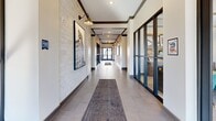 This generously proportioned hallway serves as an impressive corridor, blending modern and industrial design elements into a cohesive and welcoming passage. The space is defined by its striking exposed dark wood beam ceiling accents, which frame the length of the corridor and add architectural depth and character. One side of the hallway features a textured light-toned brick wall, providing a warm, tactile contrast to the smooth white walls on the opposite side. Large-format neutral tile flooring runs the full length of the corridor, lending a clean and polished aesthetic, while a runner rug anchors the central walkway and adds a practical touch. Wall-mounted industrial-style sconce lighting punctuates the space at intervals, providing both functional illumination and stylish detail. Framed artwork — including bold black-and-white architectural photography — adorns the walls, adding a curated, professional character to the environment. Floor-to-ceiling dark-framed glass partition walls line portions of the corridor, offering visual connectivity to adjacent spaces while maintaining a sleek, contemporary look. At the far end, a set of dark-framed glass double doors opens to the outdoors, flooding the hallway with abundant natural light and offering views of the surrounding greenery. The overall ambiance is polished, professional, and thoughtfully designed.