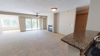 This single-floor unit is designed with an open-concept layout that connects the living, dining, and kitchen areas into one cohesive, functional space. The living room is generously proportioned, featuring plush neutral carpeting, a ceiling fan, and a glass-front fireplace that serves as a central focal point. A sliding glass door draws in natural light and provides views of surrounding greenery, enhancing the airy feel of the space.

The kitchen is well-appointed with warm-toned wood cabinetry extending to the ceiling, dark granite countertops, and black appliances including a range, microwave, refrigerator, and dishwasher. A peninsula with a double basin sink and matching granite surface separates the kitchen from the living area, offering both additional workspace and an open sightline throughout the unit.

A bedroom features carpet flooring, neutral walls, and a window with blinds that allows natural light to filter in. The bathroom includes tile flooring in an earthy stone pattern, a granite-topped vanity with ample cabinet storage, a large mirror, and a full bathtub. Wood-paneled interior doors and consistent trim throughout contribute to the cohesive, polished aesthetic of this well-maintained space.