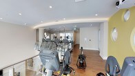 This single-floor fitness center offers a clean, contemporary design with a thoughtfully open layout that promotes an energizing workout environment. The space features warm hardwood flooring throughout, complemented by bright white walls and a smooth ceiling lined with recessed lighting and ceiling fans that ensure consistent airflow and comfort during exercise.

The facility is well-stocked with a variety of cardio equipment, including Matrix treadmills, stationary and recumbent bikes, ellipticals, and a rowing machine, catering to a wide range of fitness preferences and training needs. Equipment is arranged to maximize floor space and movement flow, with glass railings along one section adding a modern architectural touch while maintaining an open sightline across the room.

Natural light filters in through windows at the far end of the space, contributing to a bright and motivating atmosphere. An emergency exit is clearly marked, and a control panel is mounted on the wall, suggesting smart building integration. The overall ambiance is polished and functional, striking a balance between a professional gym setting and an inviting, accessible environment suitable for everyday use.